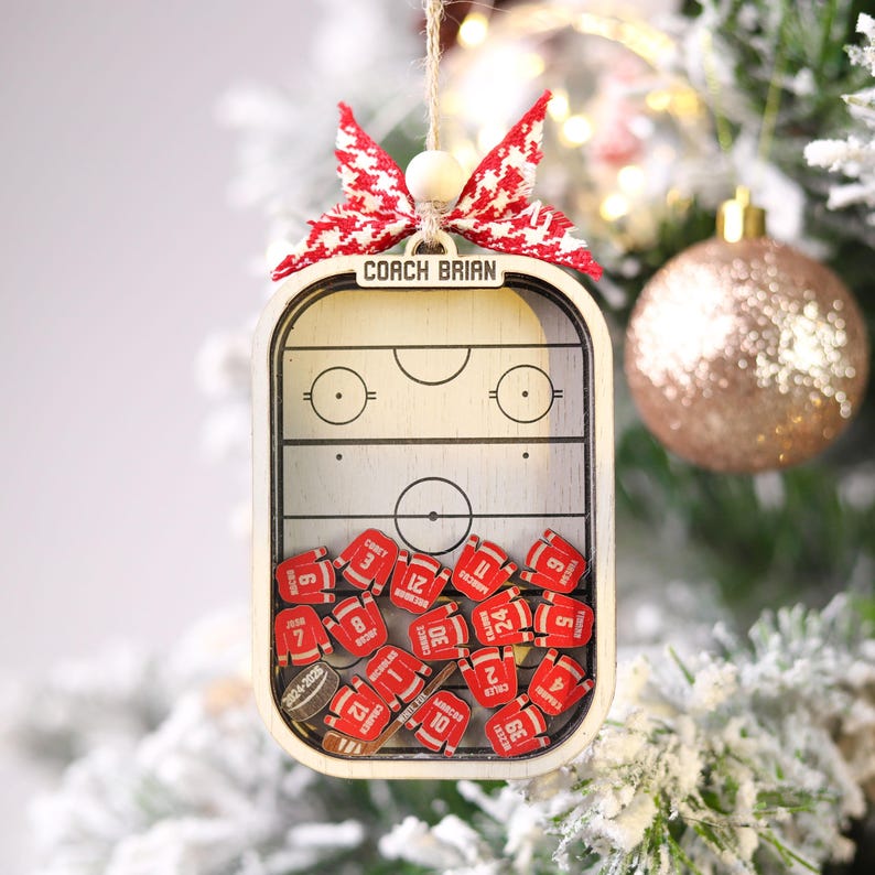 May include: Wooden hockey-themed Christmas ornament. The ornament features a hockey rink design with the text "COACH BRIAN" at the top. Inside are miniature red hockey jerseys with numbers. A red and white bow and a wooden bead are at the top.