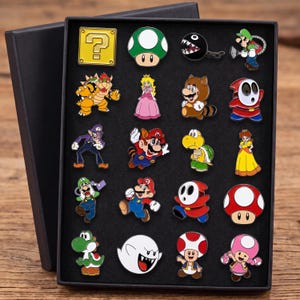 Könnte beinhalten: Eine schwarze Präsentationsbox mit zwanzig Emaille-Anstecknadeln, die verschiedene Super Mario-Charaktere und Gegenstände zeigen, darunter Mario, Luigi, Prinzessin Peach, Bowser, Yoshi, Toad, ein Fragezeichen-Block, Pilze und weitere ikonische Figuren.