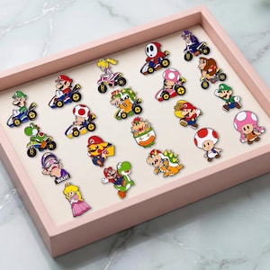May include: A collection of twenty-one colourful enamel pins featuring various video game characters from the Mario franchise, including figures in karts, motorcycles, and standing poses, all arranged neatly inside a light pink rectangular display tray.