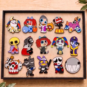 May include: A collection of fifteen colorful enamel pins arranged in a rectangular display tray. The pins feature various cartoon-style characters, including jesters, creatures with large eyes, and abstract shapes in vibrant red, blue, purple, and yellow.