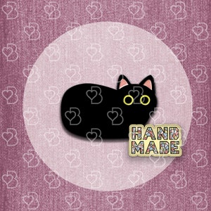 May include: Black cat pin with yellow eyes, lying down on a white background. The pin has a mosaic-style "HAND MADE" label.