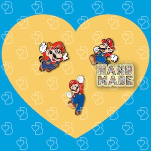 K&ouml;nnte beinhalten: Drei Emaille-Pins mit Figuren aus der Super Mario Bros. Videospielserie. Die Pins befinden sich auf einem gelben herzf&ouml;rmigen Hintergrund. Die Pins zeigen Mario, Luigi und einen Waschb&auml;r-Mario. Der Text "HAND MADE" befindet sich auf einem bunten Mosaik-Hintergrund.