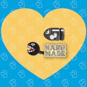 K&ouml;nnte beinhalten: Zwei Emaille-Pins auf einem gelben Herz-Hintergrund. Ein Pin ist schwarz und hat die Form eines Videospielcharakters. Der andere Pin ist mehrfarbig und sagt "HANDMADE".