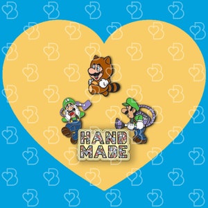 K&ouml;nnte beinhalten: Drei Emaille-Pins mit Figuren aus der Super Mario Bros. Videospielserie. Die Pins befinden sich auf einem gelben herzf&ouml;rmigen Hintergrund. Die Pins zeigen Mario, Luigi und einen Tanooki Mario. Der Text "HANDMADE" befindet sich in der Mitte des Bildes.