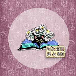 May include: Black cat peeking out from an open book with colorful flowers. The book is blue and green with a white background. The text "HAND MADE" is on a colorful mosaic background.