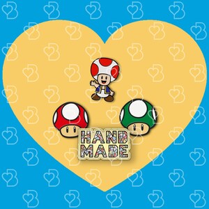 K&ouml;nnte beinhalten: Drei handgefertigte Emaille-Pins mit Figuren aus der Super Mario Bros. Videospielserie. Die Pins sind in Form eines roten und wei&szlig;en Pilzes, eines gr&uuml;nen und wei&szlig;en Pilzes und einer roten und blauen Pilzfigur mit einem blauen und wei&szlig;en Hut. Die Pins befinden sich auf einem gelben Hintergrund mit einem Herzmuster und dem Wort "HANDMADE" in bunten Mosaikfliesen.