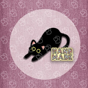 May include: Black enamel pin of a cat with yellow eyes, lying down on a white background. The pin says "HANDMADE" in colorful mosaic letters.