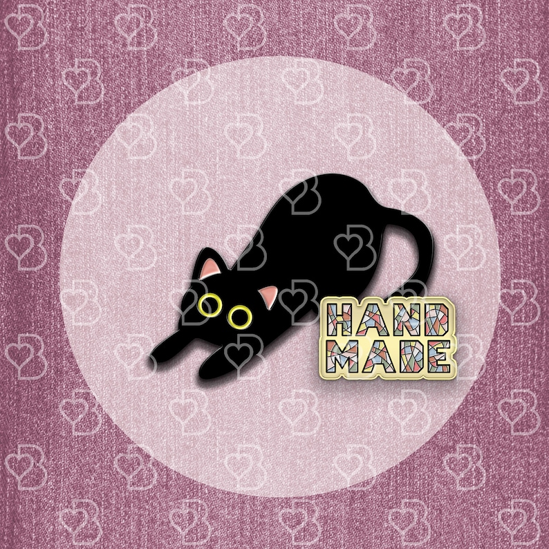 May include: Black enamel pin of a cat with yellow eyes, lying down on a white background. The pin says "HANDMADE" in colorful mosaic letters.