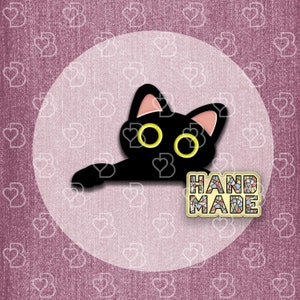 May include: Black cat with yellow eyes peeking out from behind a handmade sign that says "HAND MADE".