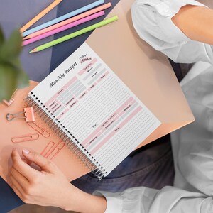 May include: A pink and white monthly budget planner with a spiral binding. The planner has sections for income, expenses, and savings. The text "Today's Expenses" is written in pink at the top of the page.
