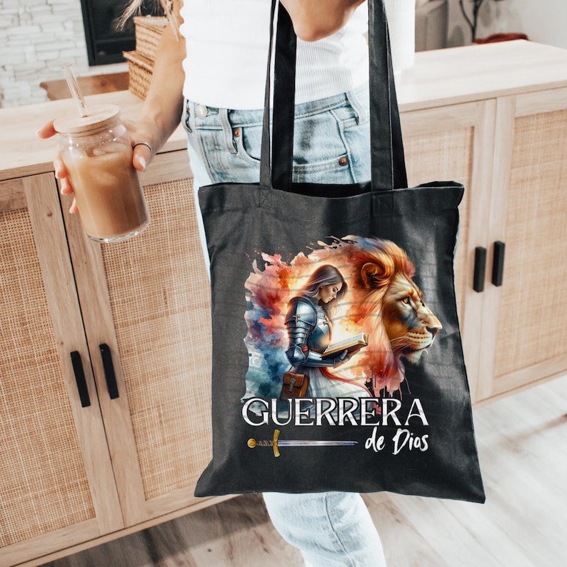 May include: Black tote bag with a watercolor design of a woman in armor holding a book and a lion's head. The text "GUERRERA de Dios" is printed on the bag.