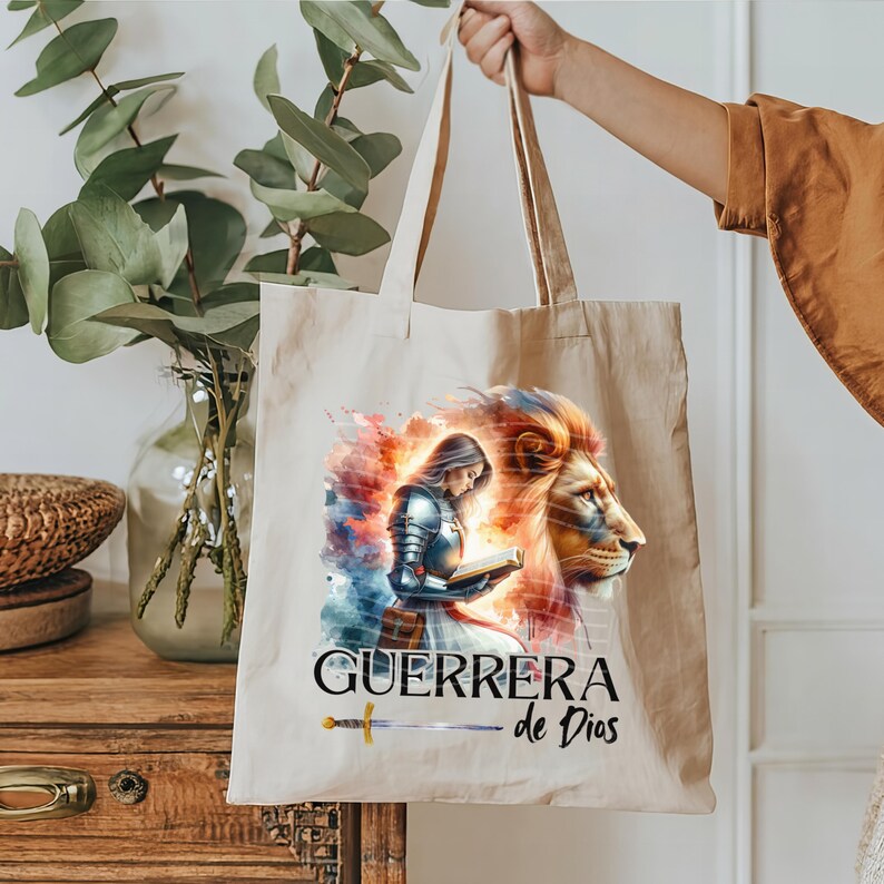 May include: A beige tote bag with a watercolor design of a woman in armor holding a book and a lion. The text "GUERRERA de Dios" is printed on the bag.