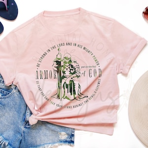 Puede incluir: Camiseta rosa con un gr&aacute;fico de un caballero con armadura y una espada. El gr&aacute;fico est&aacute; rodeado de un c&iacute;rculo con el texto "BE STRONG IN THE LORD AND IN HIS MIGHTY POWER. ARMOR OF GOD. SO THAT YOU CAN TAKE YOUR STAND AGAINST THE DEVIL'S SCHEMES."