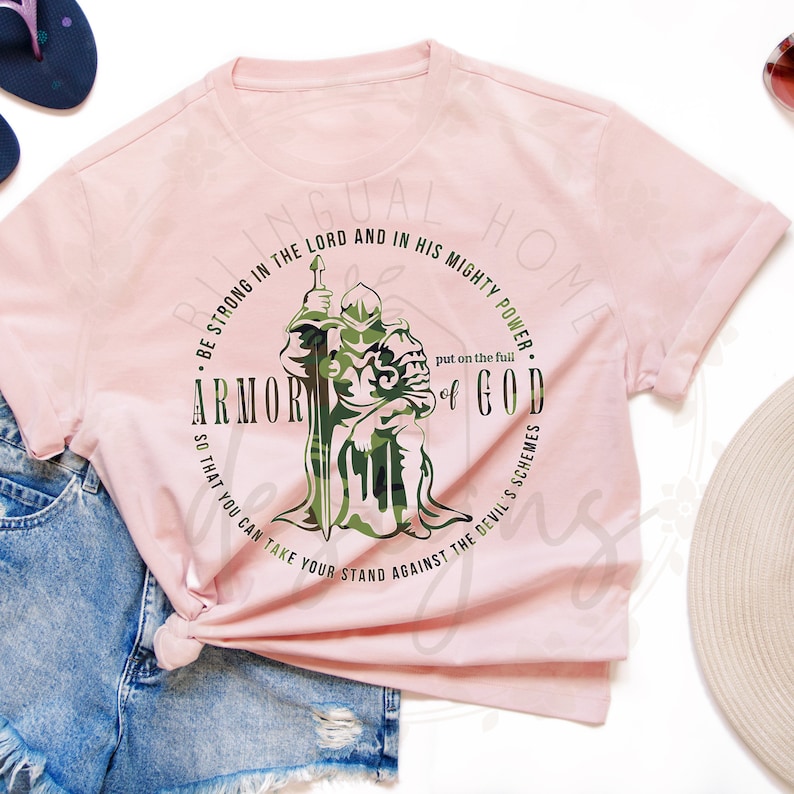 Puede incluir: Camiseta rosa con un gr&aacute;fico de un caballero con armadura y una espada. El gr&aacute;fico est&aacute; rodeado de un c&iacute;rculo con el texto "BE STRONG IN THE LORD AND IN HIS MIGHTY POWER. ARMOR OF GOD. SO THAT YOU CAN TAKE YOUR STAND AGAINST THE DEVIL'S SCHEMES."