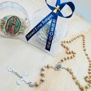 May include: A wooden rosary with a silver cross and a small round image of the Virgin Mary. The rosary is in a clear plastic bag with a blue ribbon that says "Manuel James Rodriguez 08-20-84 05-18-24".