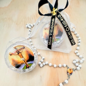 May include: White rosary beads with a silver cross and a small round image of Jesus on the cross. The rosary is in a clear plastic container with a black ribbon and a tag that reads "Mario Villalpando 10-14-1979  04-07-2024".