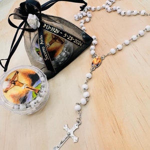 May include: White rosary beads with a silver crucifix and a small image of Jesus. The rosary is in a black organza bag with a gold ribbon that reads "Socorro Avila 7-13-1929 - 5-25-2021".