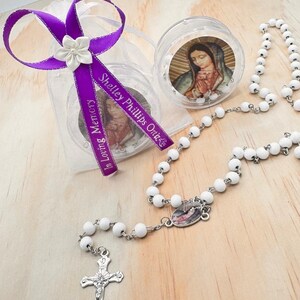 May include: White rosary beads with a silver crucifix and a small round container with a religious image. The container is in a clear plastic bag with a purple ribbon that says "Shelley Phillips Ortiz" and "In Loving Memory".