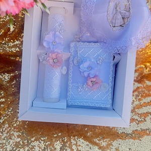May include: A white baptism set in a white box, including a decorated candle with pink and lavender flowers, a small book, and a lace-trimmed bonnet with a religious image. The set is presented on a gold sequined surface.