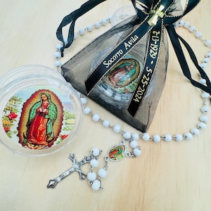 May include: A white rosary with a silver cross and a small silver charm featuring an image of the Virgin Mary. The rosary is displayed with a small glass jar containing a picture of the Virgin Mary and a black organza bag with a black ribbon that reads "Socorro Avila 7-13-1929 - 5-25-2024".