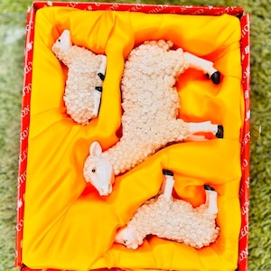 May include: Three white ceramic sheep figurines in a red and yellow box. The sheep are in various poses, including lying down and standing up.