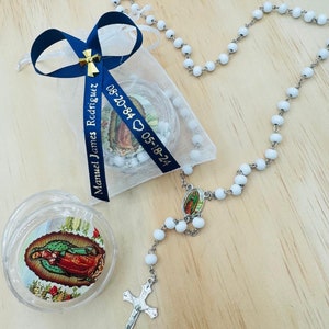 May include: White rosary beads with a silver cross and a small silver charm featuring a religious image. The rosary is packaged in a white organza bag with a blue ribbon and a personalized tag that reads "Manuel James Rodriguez 08-20-84  05-18-24".