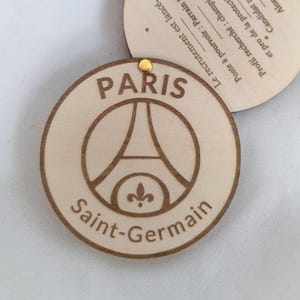 Puede incluir: Un colgante circular de madera con el logotipo del Paris Saint-Germain, que incluye la Torre Eiffel y la flor de lis. Las palabras "PARIS" y "Saint-Germain" est&aacute;n grabadas en marr&oacute;n oscuro. Una peque&ntilde;a pieza met&aacute;lica dorada est&aacute; en la parte superior. Texto en franc&eacute;s.