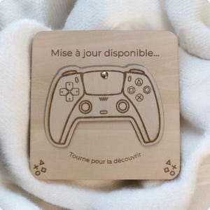 Peut inclure: Puzzle en bois en forme de manette de jeu, avec des détails gravés. Les textes "Mise à jour disponible..." et "Tourne pour la découvrir" sont visibles. Le puzzle est en bois clair et comporte une petite attache métallique.