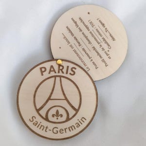 Puede incluir: Dos posavasos redondos de madera. Uno presenta el logotipo del Paris Saint-Germain, con la Torre Eiffel y la flor de lis, y las palabras "Paris" y "Saint-Germain". El segundo posavasos tiene texto en franc&eacute;s.