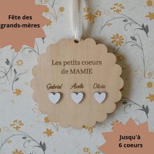 Peut inclure: Plaque en bois en forme de cœur avec des bords festonnés, portant l'inscription "Les petits coeurs de MAMIE" et les noms Gabriel, Axelle et Olivia. Trois cœurs blancs ornent le bas. Un ruban blanc permet de suspendre la plaque. Le texte "Fête des grands-mères" et "Jusqu'à 6 coeurs" sont également présents.