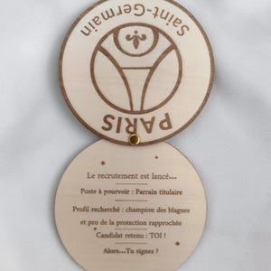 Puede incluir: Disco de madera de dos partes con las palabras "Saint-Germain" y "Paris" grabadas en la parte superior. La parte inferior tiene texto en franc&eacute;s, incluyendo "Le recrutement est lanc&eacute;..." y "Alors...Tu signes ?"