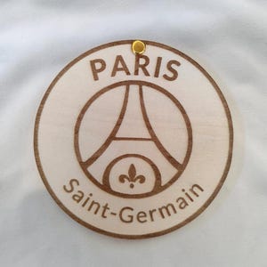 Puede incluir: Adorno redondo de madera con el logotipo del Paris Saint-Germain. El dise&ntilde;o, grabado en marr&oacute;n, muestra la Torre Eiffel y las palabras "PARIS" y "Saint-Germain". Un peque&ntilde;o ojal met&aacute;lico dorado en la parte superior.