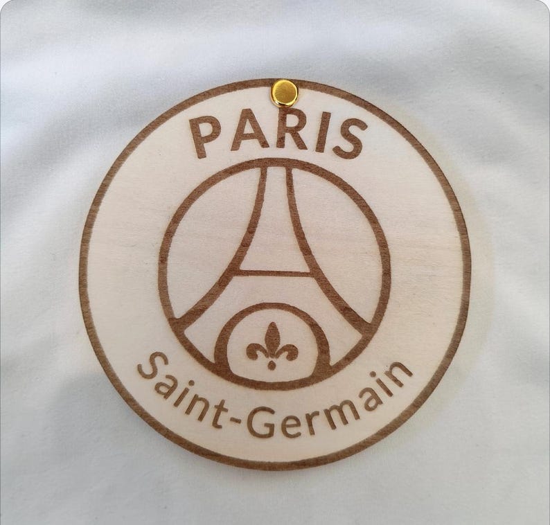 Puede incluir: Adorno redondo de madera con el logotipo del Paris Saint-Germain. El dise&ntilde;o, grabado en marr&oacute;n, muestra la Torre Eiffel y las palabras "PARIS" y "Saint-Germain". Un peque&ntilde;o ojal met&aacute;lico dorado en la parte superior.