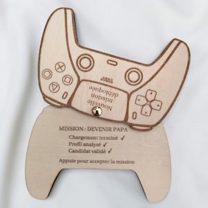Op de afbeelding: Houten plaquette in de vorm van een gamecontroller, met gegraveerde details. Het bovenste deel heeft knoppen en een directioneel pad, met tekst. Het onderste deel draagt de tekst "MISSION: DEVENIR PAPA".