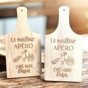 Peut inclure: Deux planches à découper en bois clair avec des poignées. Chaque planche porte l'inscription gravée "Le meilleur APÉRO c'est avec Papa" et "Le meilleur APÉRO c'est avec Papi" avec des illustrations de collations et de cocktails.