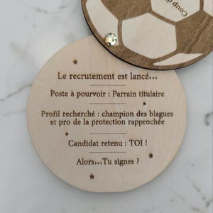 Peut inclure: Deux dessous de verre ronds en bois. L'un présente un motif de ballon de football, l'autre contient du texte en français : "Le recrutement est lancé... Poste à pourvoir: Parrain titulaire... Candidat retenu: TOI! Alors... Tu signes ?" Une petite gemme est fixée au dessous de verre.