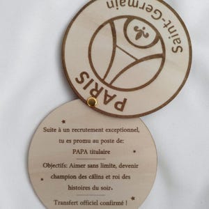 Peut inclure: Un disque en bois avec les mots "Paris Saint-Germain" et un logo gravé sur la moitié supérieure. La moitié inférieure révèle le texte : "Suite à un recrutement exceptionnel, tu es promu au poste de: PAPA titulaire."