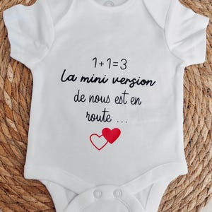 Peut inclure: Body de bébé blanc à manches courtes. Le body porte l'inscription "1+1=3 La mini version de nous est en route ..." en écriture noire, avec deux cœurs rouges. Fabriqué en matière douce, il possède des boutons-pression.