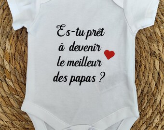 May include: White baby bodysuit featuring the French text "Es-tu prêt à devenir le meilleur des papas ?" in black script, with a small red heart. The garment is presented on a woven, light brown surface.