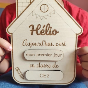 Peut inclure: Une plaque en bois clair en forme de maison avec des gravures laser indiquant Hélio, Aujourd'hui, c'est, mon premier jour, en classe de, et CE2. La plaque présente une cheminée, une fenêtre circulaire et une illustration de crayon.