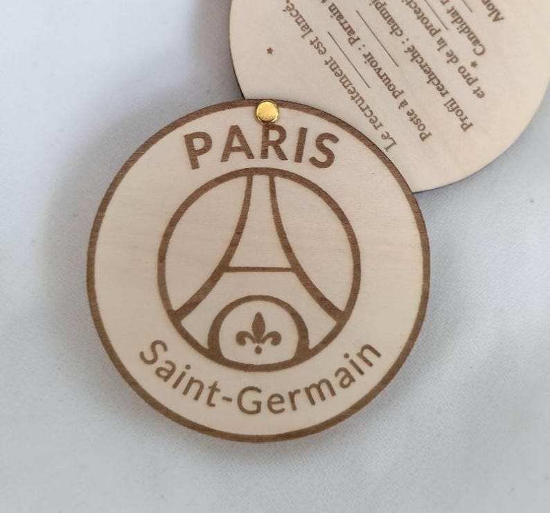 Puede incluir: Un colgante circular de madera con el logotipo del Paris Saint-Germain, que incluye la Torre Eiffel y la flor de lis. Las palabras "PARIS" y "Saint-Germain" est&aacute;n grabadas en marr&oacute;n oscuro. Una peque&ntilde;a pieza met&aacute;lica dorada est&aacute; en la parte superior. Texto en franc&eacute;s.