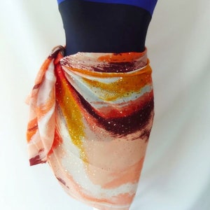 May include: A colourful abstract print sarong with orange, red, and gold accents. The sarong is tied around a white mannequin wearing a blue and black swimsuit.