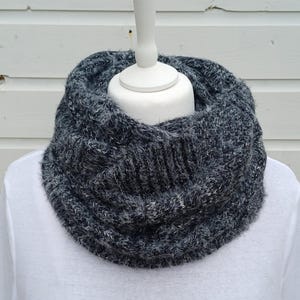 May include: A dark grey and white speckled infinity scarf with a chunky knit texture. The scarf features a thick, fuzzy yarn composition with a ribbed pattern throughout, creating a voluminous, circular loop design that sits around the neck.