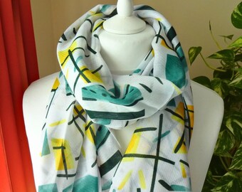 May include: A white scarf featuring a geometric pattern of teal green rectangles, yellow rectangular blocks, and thin black intersecting lines. The lightweight fabric is draped around a white mannequin neck, displaying the abstract print clearly.
