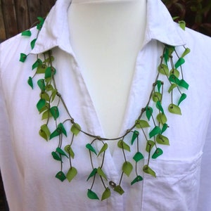 May include: A necklace featuring multiple thin brown cords adorned with numerous small, translucent green and lime-coloured leaf-shaped pendants. The leaves are attached at varying intervals along the cords, creating a layered, cascading effect.