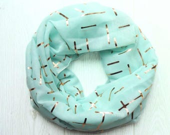 May include: A light mint green infinity scarf featuring a repeating pattern of metallic copper-coloured horizontal dashes and cross symbols. The lightweight fabric is arranged in a circular loop, showcasing the shiny foil print across the surface.