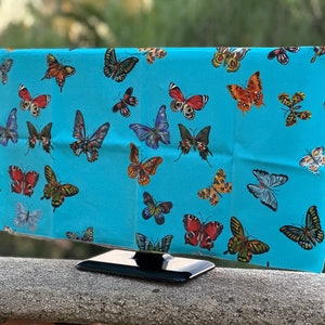 May include: A turquoise fabric with a repeating pattern of colorful butterflies. The fabric is draped over a black television stand.
