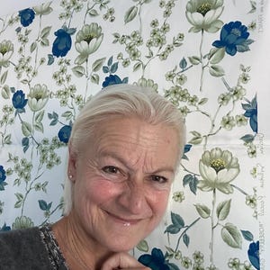 May include: A woman with short, white hair smiles at the camera. She is wearing a gray sweater and has her hand on her chin. The background is a white fabric with a floral pattern in shades of blue and green.