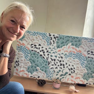 Op de afbeelding: Een TV-schermbeveiliging met een bloemenpatroon in tinten teal, roze, wit en zwart. De beschermer staat op een standaard met een dennenappel, een kaars en een decoratief object op een houten oppervlak.