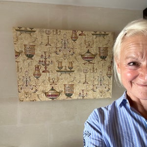 May include: A close-up of a woman with blonde hair smiling at the camera. She is wearing a blue and white striped shirt. Behind her is a wall with a beige and gold wallpaper featuring a repeating pattern of ornate vases and candlesticks.